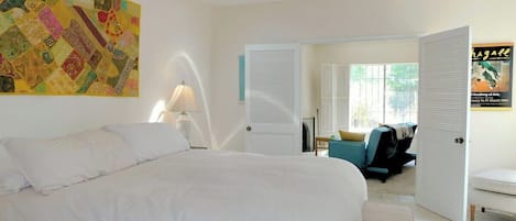 Quarto de hotel com uma cama grande, uma obra de arte colorida na parede, uma porta que dá para uma varanda e um cartaz na parede.