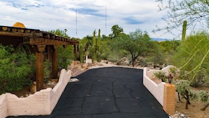 A paved pathway leading to a wooden structure with a roof, surrounded by cacti and desert vegetation.
