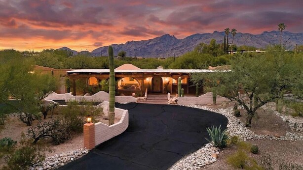 Una casa de estilo desértico con un camino pavimentado, rodeada de cactus y arbustos, y una cadena montañosa al fondo.