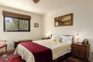 A bedroom with a bed, bedside tables, a nightstand, a window with a view, and a painting on the wall.
