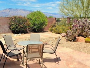 An outdoor patio with a round table and four chairs, surrounded by desert vegetation and a view of mountains.