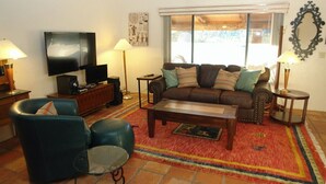 Ein Wohnzimmer mit einer Couch, Sesseln, einem Couchtisch, einem Fernseher und einem großen Fenster.