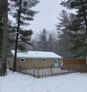 Una casa con tejado cubierto de nieve, rodeada por una cerca de madera y altos árboles de hoja perenne.