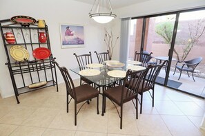 A dining area with a glass table surrounded by chairs, a metal rack with dishes, and a large window with a view of the outdoors.