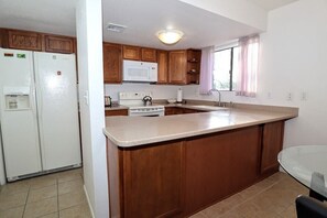 A kitchen with wooden cabinets, a white refrigerator, a microwave, a stove, and a glass dining table. - Ventana Vista 1179 Condo (Tucson)