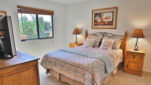 Une chambre à coucher avec un lit, deux tables de chevet, une télévision, une fenêtre et un tableau encadré au mur.