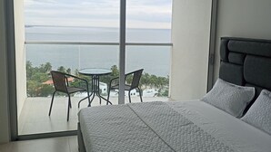 A bedroom with a large window overlooking a balcony with a table and chairs, a bed with a grey headboard, and a view of the sea and palm trees. - Ocean View Apartment w/ Private Beach & Pools Santa Marta – Pozos Colorados (Santa Marta)