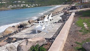 Zona frente a la playa con sillas de lounge blancas, un camino de concreto y vista al océano y una colina al fondo.