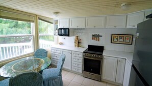 Una cocina con armarios blancos, microondas, horno y refrigerador. Hay una mesa de vidrio con sillas y una ventana con persianas.