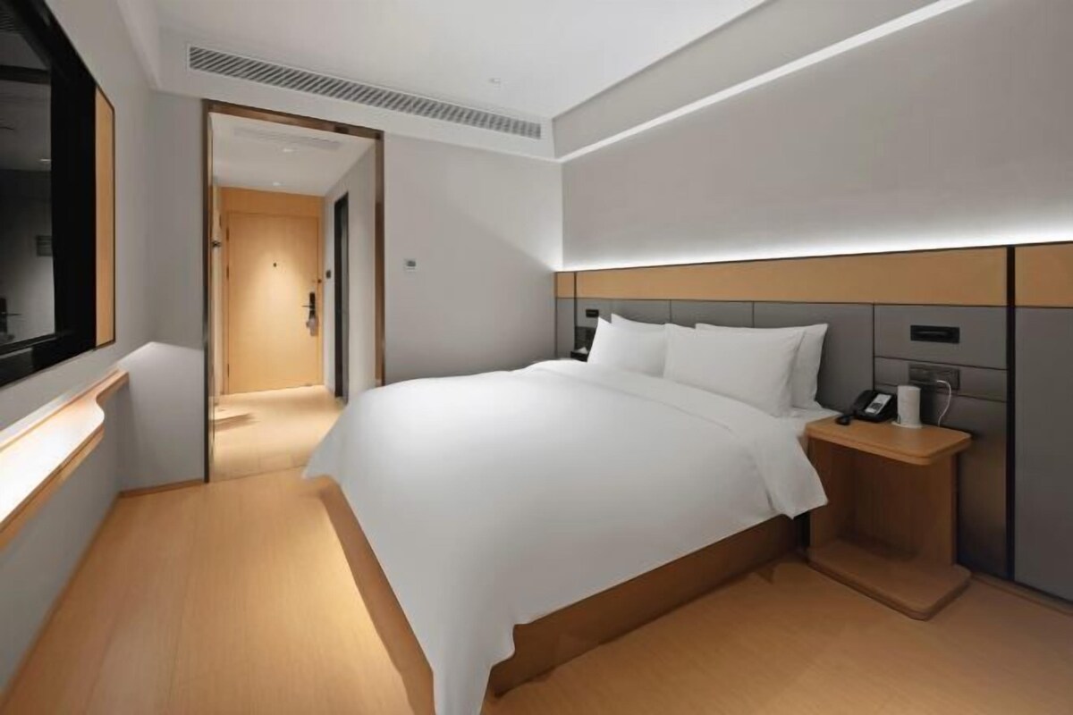 Quarto de hotel moderno com uma cama grande, mesa de cabeceira e vista através de uma porta aberta.