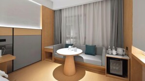 A modern hotel room with a round wooden table, a sofa with cushions, a small cabinet with a refrigerator, and a large window with curtains.
