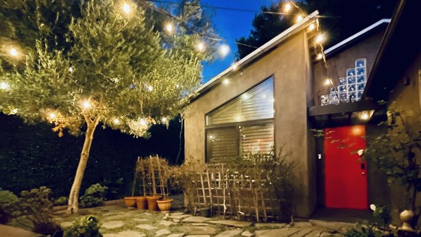 Un sendero de piedra conduce a un garaje con puerta roja, rodeado de plantas en macetas y luces colgantes.