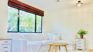 Una sala de estar moderna con un sofá seccional blanco, una mesa de centro redonda de madera, una alfombra mullida blanca, una planta en maceta y una ventana con un adorno marrón.