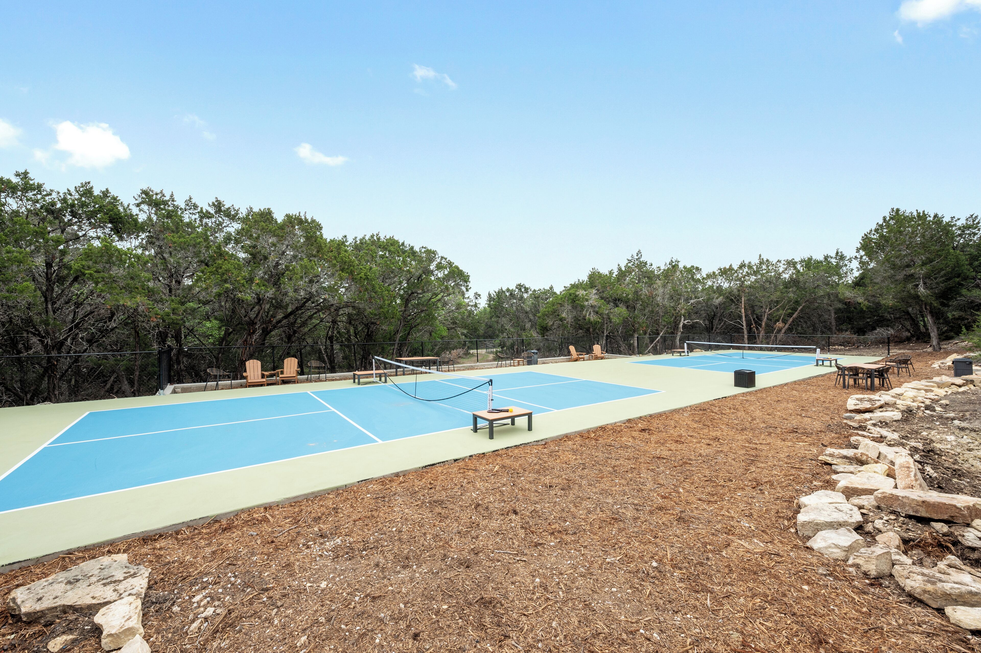 An outdoor tennis court with a net, surrounded by trees and equipped with outdoor seating.