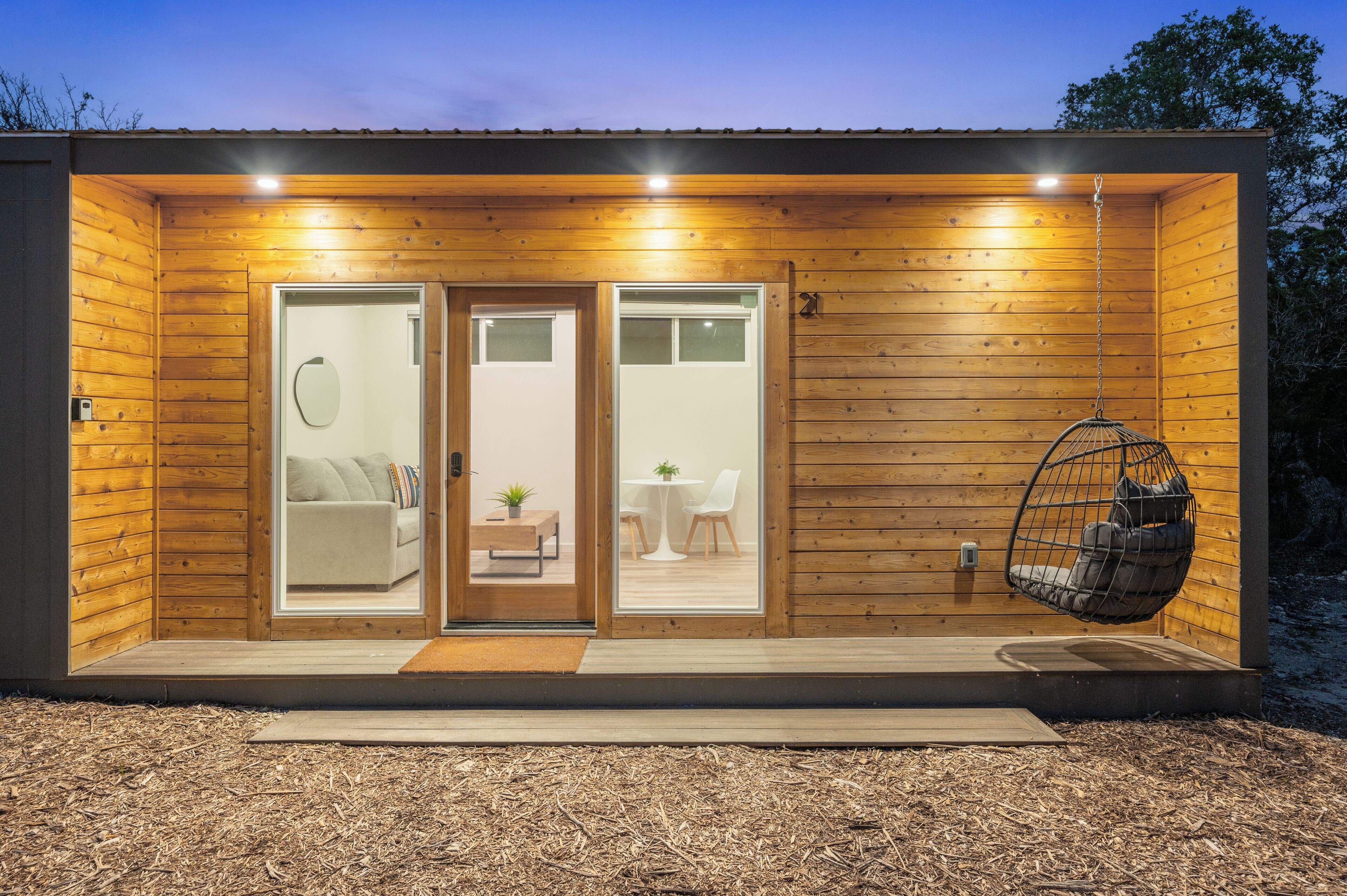 A modern wooden cabin with a front door, a small seating area with a table and chairs, and a hanging chair.