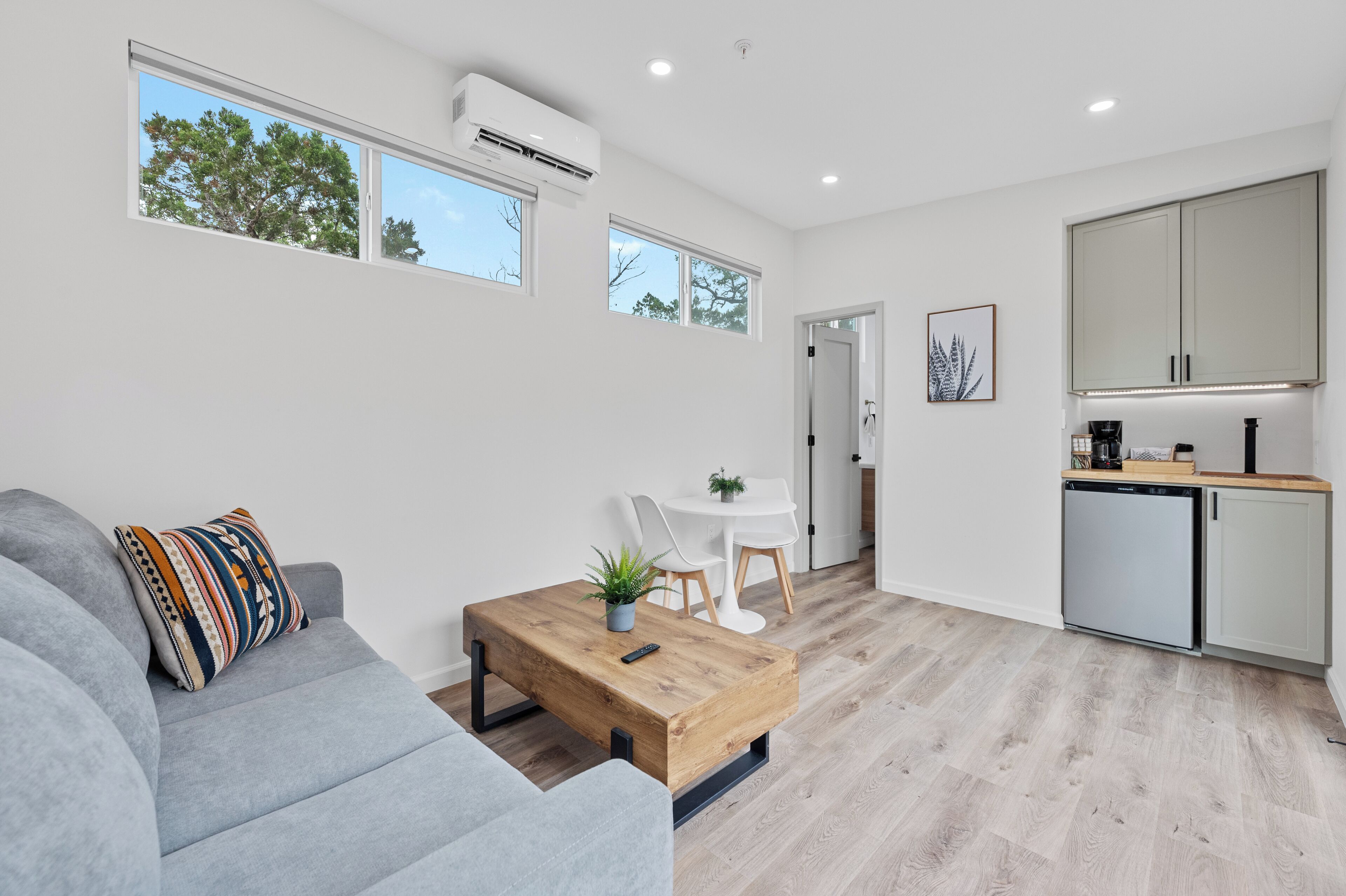 A modern living room with a grey sofa, a wooden coffee table, and a small table with chairs. There is a kitchen area with cabinets and a dishwasher.