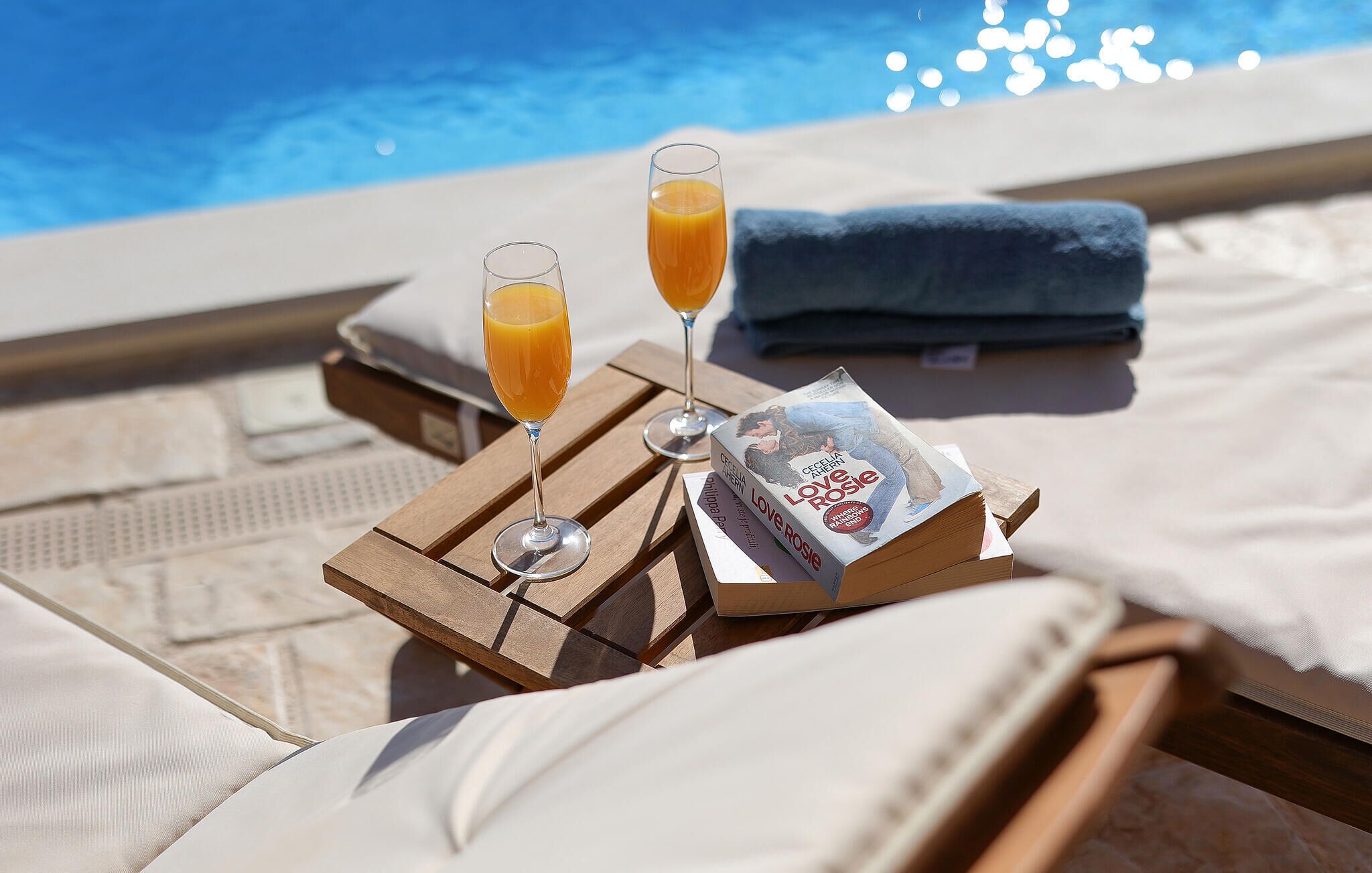 A poolside setting with a lounge chair, a towel, two glasses of orange juice, and a book titled "Love Rosie".