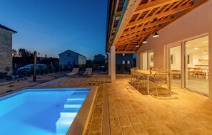 Un espace aménagé au bord de la piscine, avec un patio couvert, des sièges d’extérieur et une vue sur un bâtiment en pierre en arrière-plan.