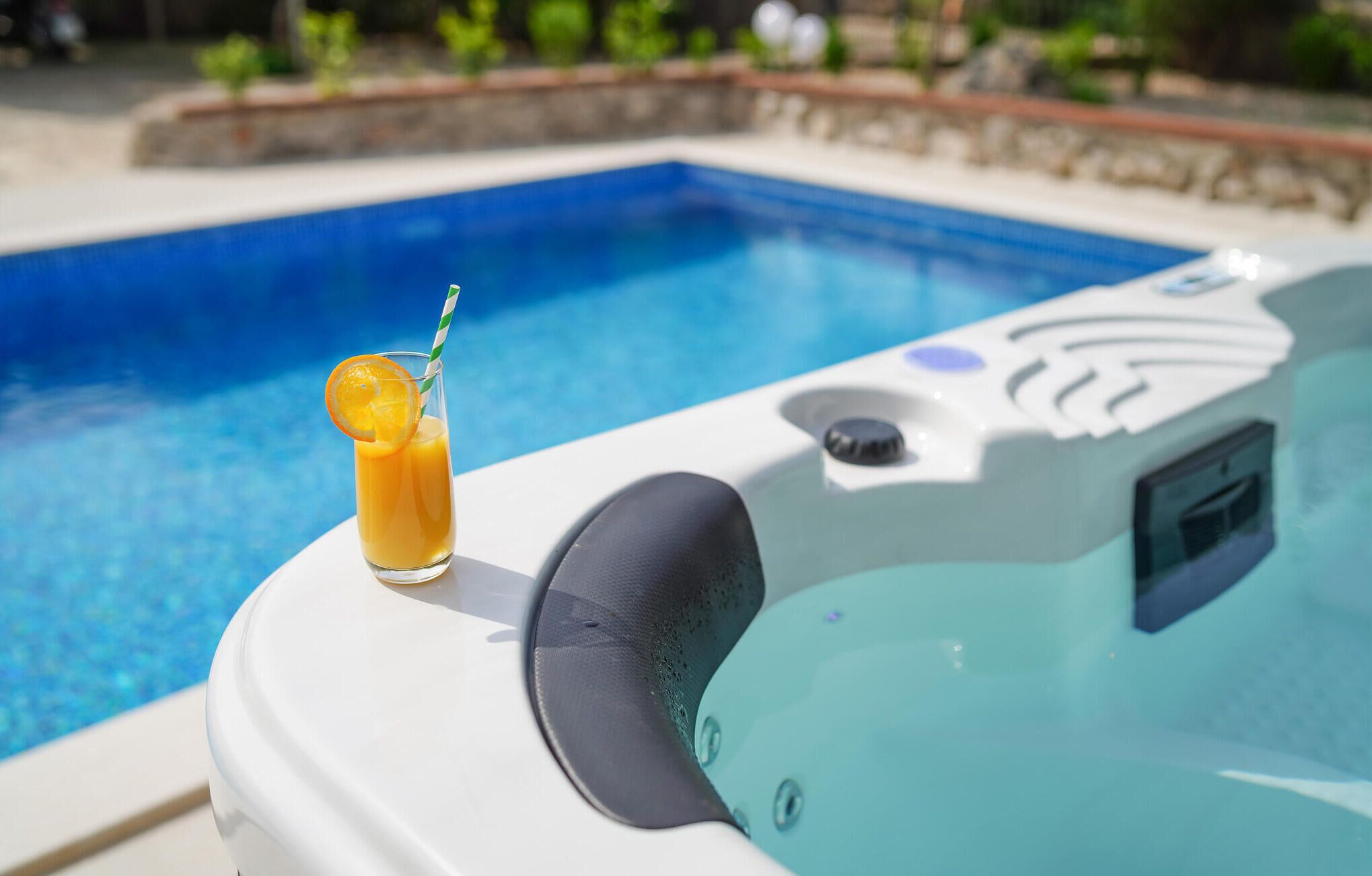 A hot tub with a glass of orange juice and a straw, next to a swimming pool.