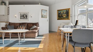 A modern living room with a brown leather sofa, a white coffee table, and a dining area with a white table and grey chairs. There are framed artworks on the walls and a large window providing natural light.
