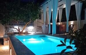 A swimming pool with a stone border, a tree, and a covered patio area with a grill.