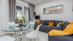 A modern living room with a glass dining table, white chairs, a grey sofa with yellow and grey cushions, and a large mirror on the wall. - Amazing home in Zdrelac (Zdrelac)