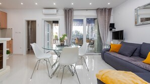 A modern living room with a glass dining table, white chairs, a grey sofa with yellow cushions, and a TV mounted on the wall. - Amazing home in Zdrelac (Zdrelac)