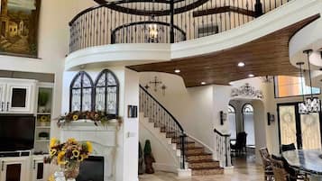 A spacious living area with a grand staircase, a fireplace with a floral arrangement, and a dining area with wicker chairs.