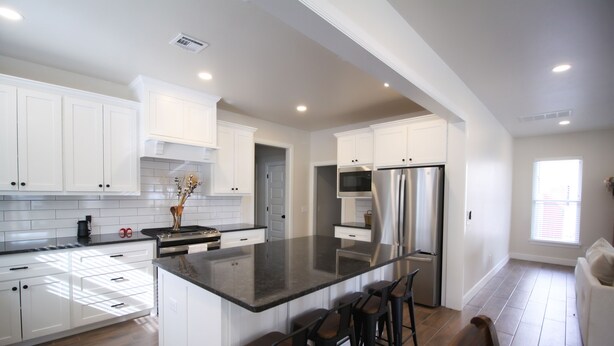 A modern kitchen with white cabinetry, a large island with bar stools, stainless steel appliances, and a view to the adjacent living area. - The Campus Cottage on Elm is the ideal University of Oklahoma gameday residence (Norman)