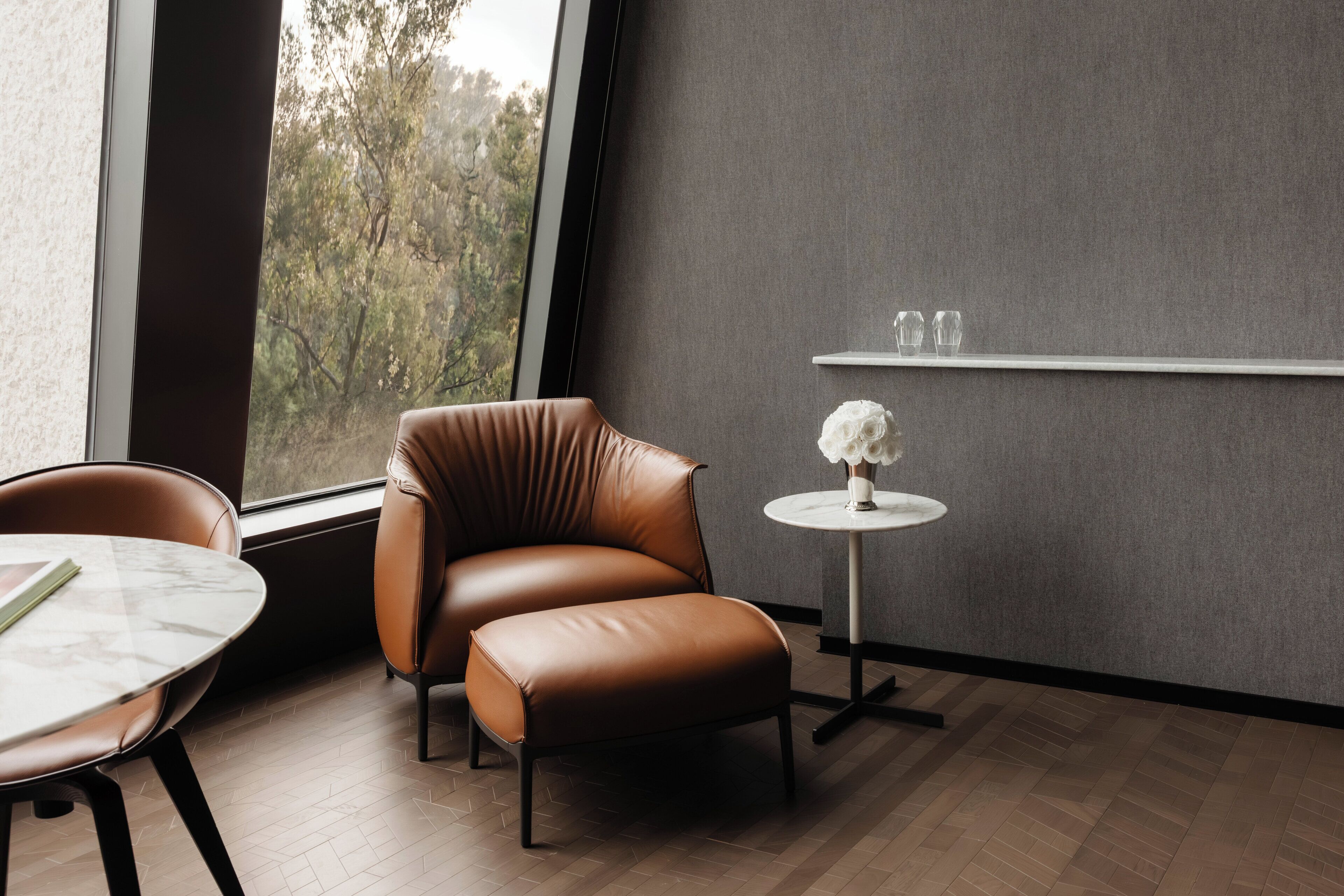 Una habitación moderna con un sillón de cuero, una mesita redonda con un florero y una ventana con vistas a árboles.