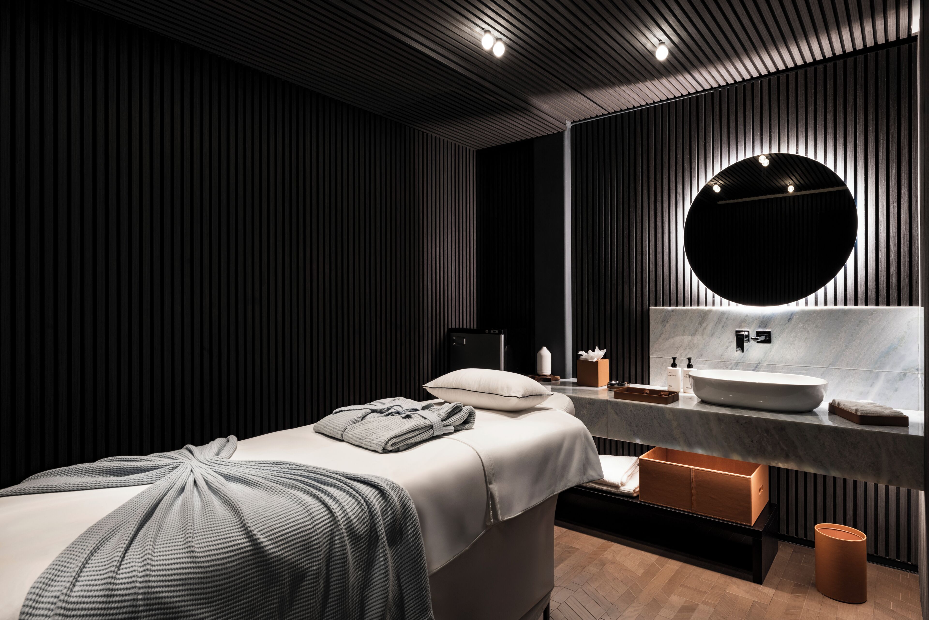 Quarto de hotel moderno com cama, espelho redondo, pia e chão de madeira.