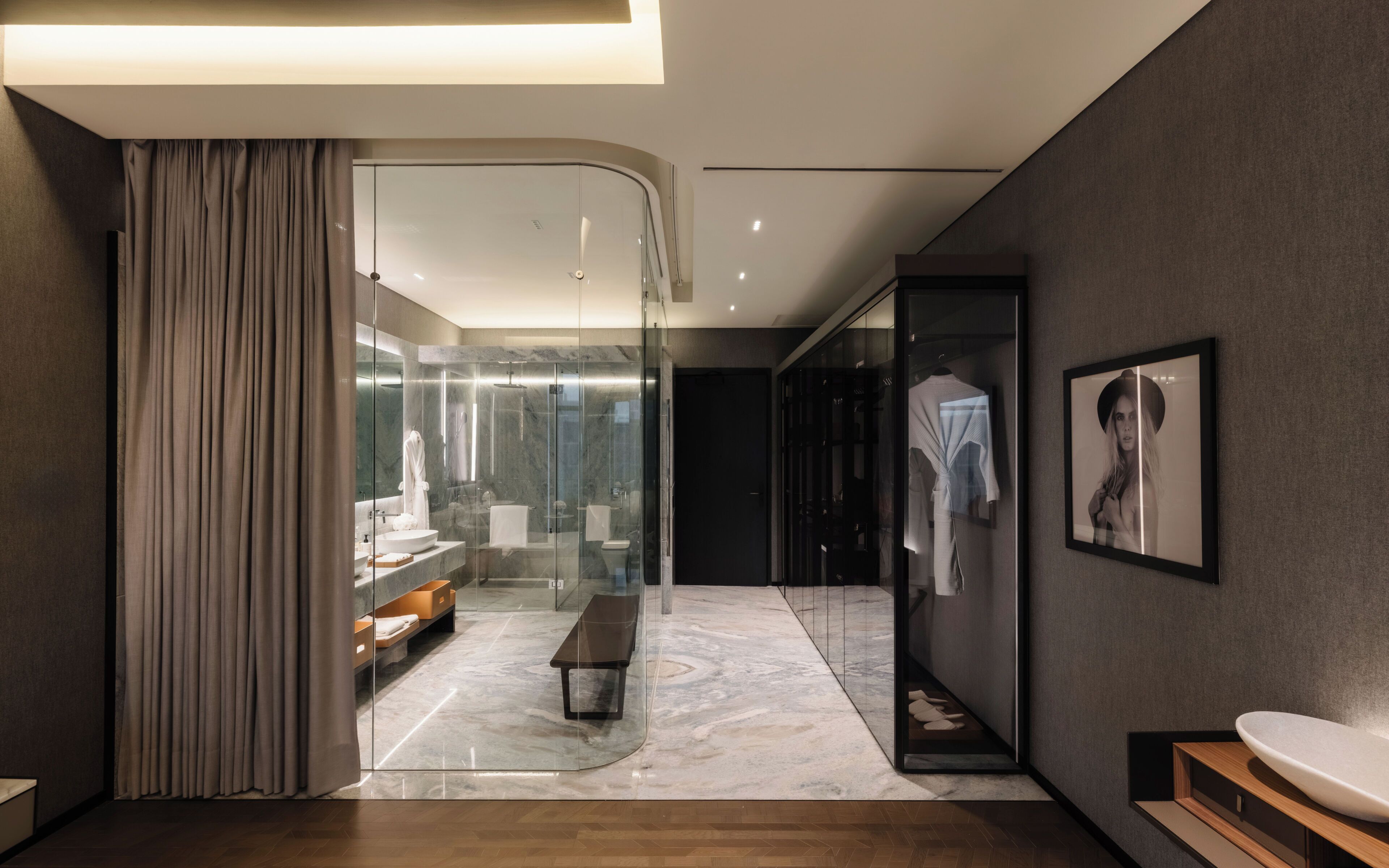 A modern hotel bathroom with a glass-enclosed shower, marble flooring, and a wooden bench.