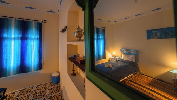 A room with a bed, a nightstand, a window with curtains, a small table, and a decorative bowl on a shelf.
