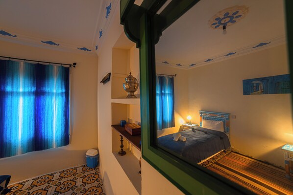 Una habitación con cama, mesita de noche, ventana con cortinas, una pequeña mesa y un cuenco decorativo en una estantería.