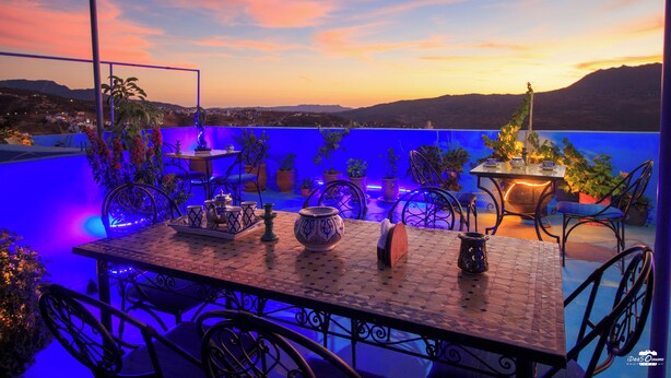 A rooftop terrace with a table set for dining, surrounded by chairs and potted plants, overlooking a cityscape at sunset. - Casa voyager with secret garden terrace (Chefchaouen av moulay)