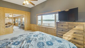 Un dormitorio con una cama grande, paredes de madera, ventilador de techo, cómoda, televisor y ventana con vista a un lago.