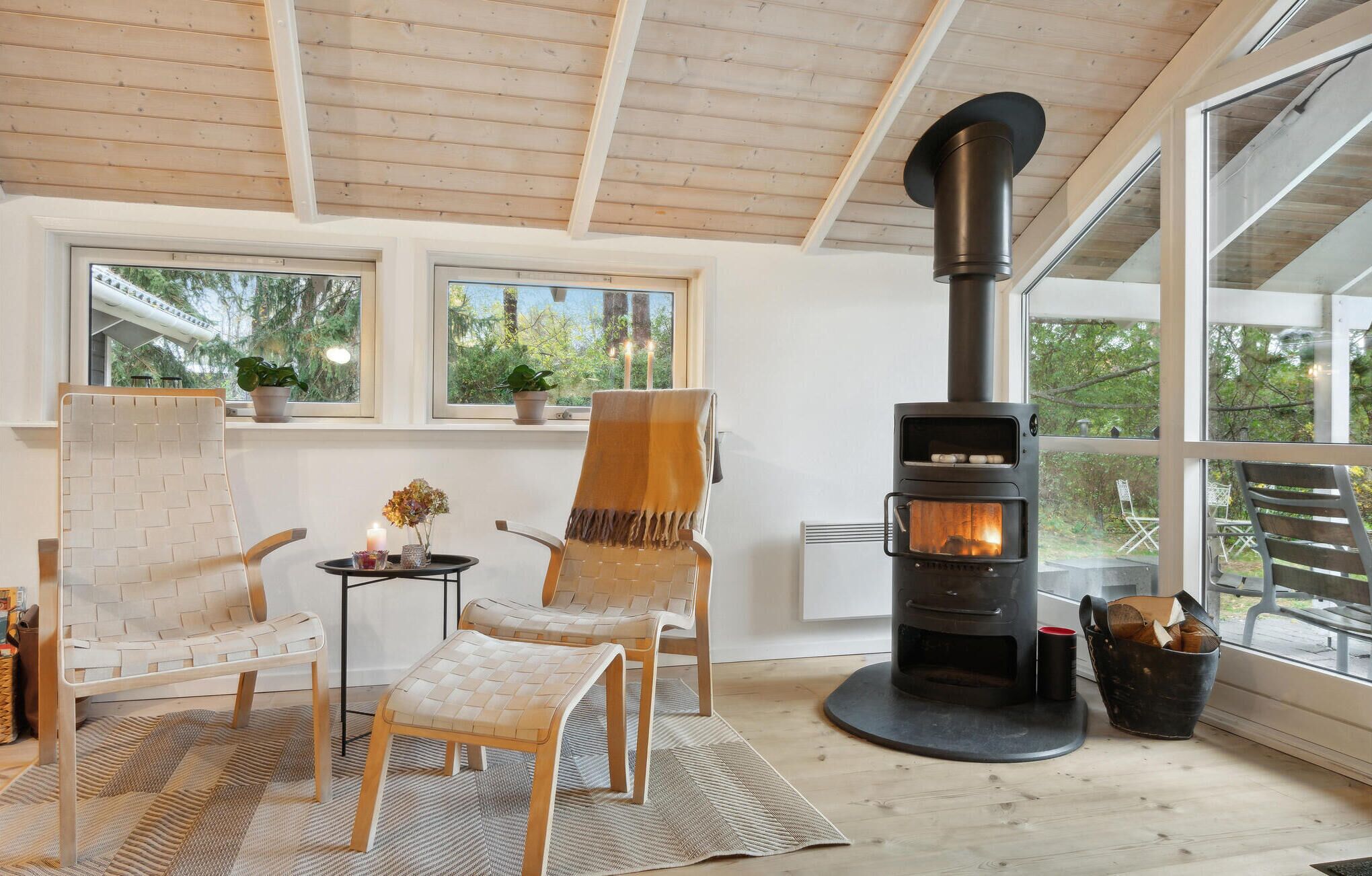 A cozy room with a wood-burning stove, two chairs, a table with candles, and large windows offering a view of trees.