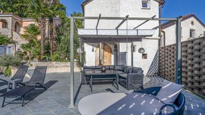 Un espace extérieur aménagé, abrité et doté de chaises longues et d’une table, ainsi qu’une pergola et un bâtiment blanc avec une porte en bois.