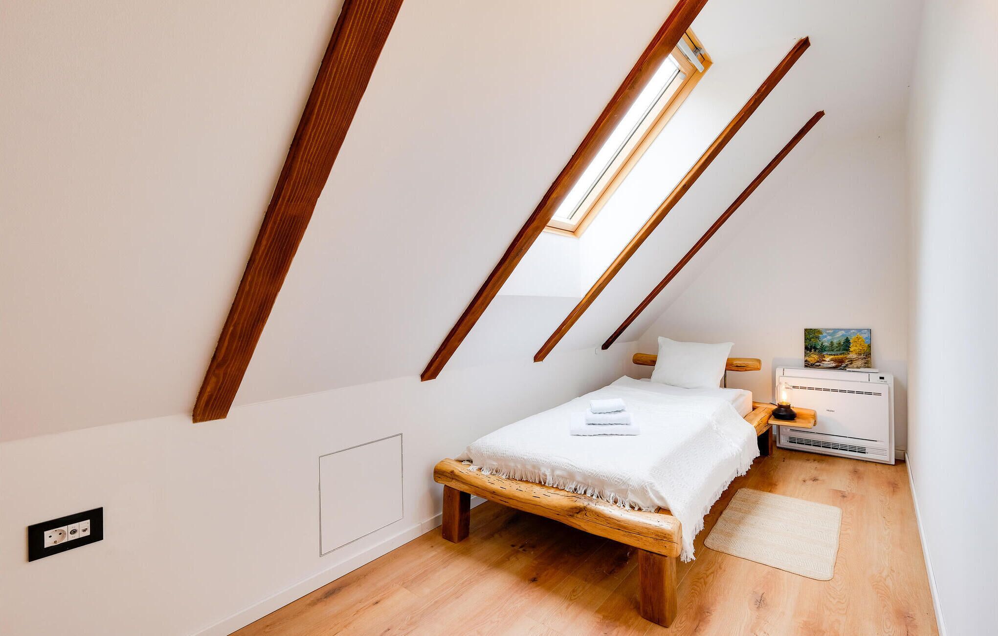 Une petite chambre bien éclairée, avec un plafond en pente, un lit simple, une table de chevet avec une lampe et un radiateur.