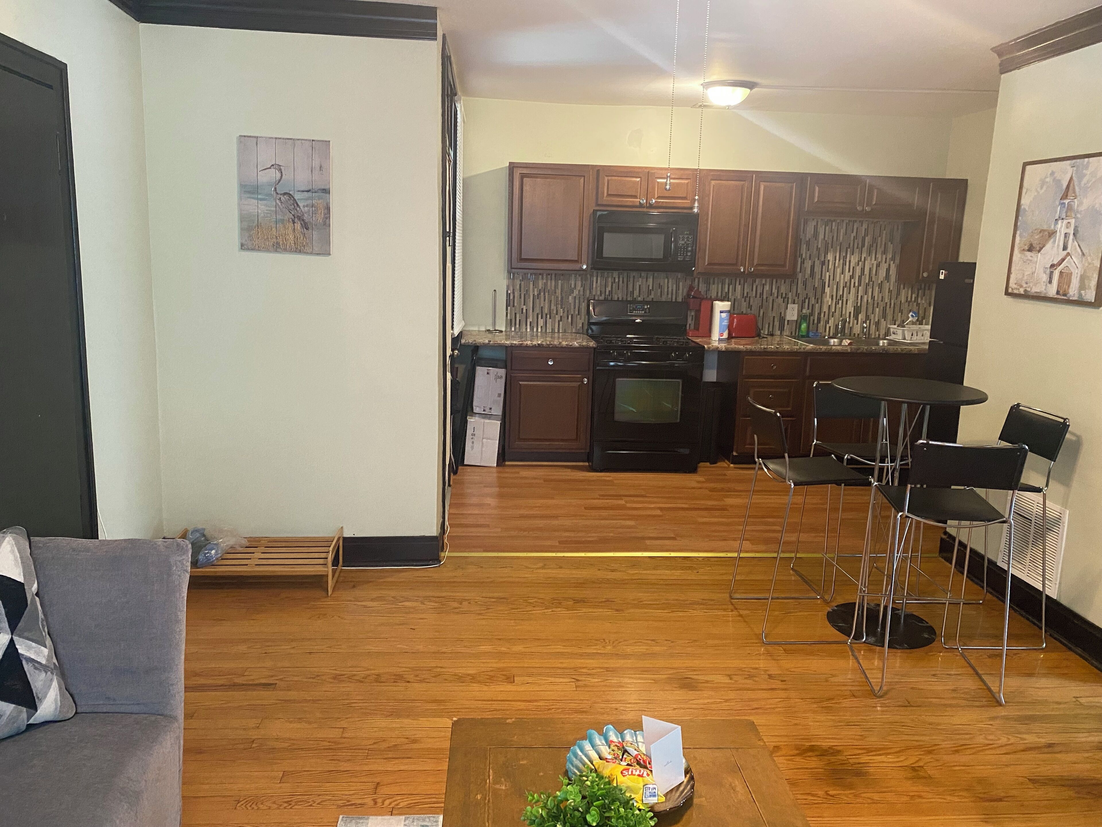 Uma sala de estar com um sofá cinza, uma mesa de centro de madeira com uma planta, e uma cozinha ao fundo com armários de madeira, um micro-ondas e um fogão.
