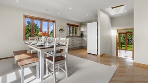 A modern kitchen with white cabinetry, a dining area with a wooden table and chairs, and large windows offering a view of greenery. - Beachside Luxury - Laingholm Holiday Apartment (Auckland)