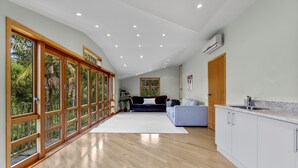 A spacious living area with a large sliding glass door leading to a balcony, a kitchen with white cabinets, and a ceiling with recessed lighting. - Beachside Luxury - Laingholm Holiday Apartment (Auckland)