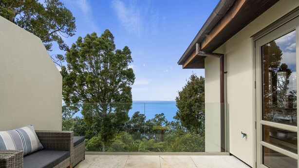 A balcony with a view of the sea, a wicker sofa, and a glass door. - Beachside Luxury - Laingholm Holiday Apartment (Auckland)