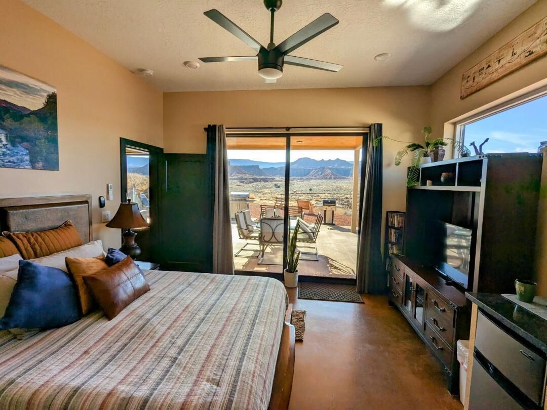 Un dormitorio con cama, ventilador de techo, televisión, escritorio y vista al desierto a través de una puerta corrediza.