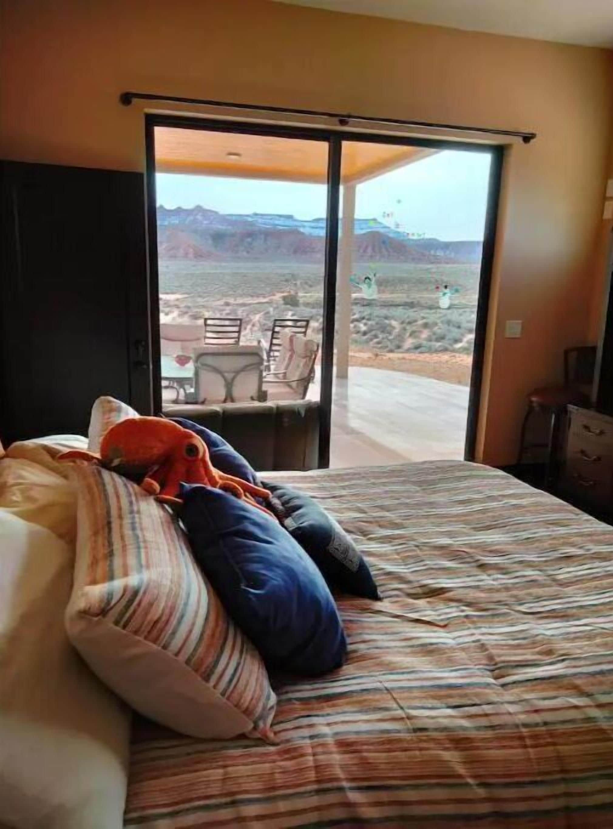 A bedroom with a bed, pillows, a striped blanket, and a view of a desert landscape through an open sliding door.