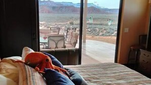 Un dormitorio con cama, almohadas, una manta a rayas y vistas a un paisaje desértico a través de una puerta corrediza abierta.