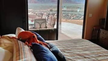 A bedroom with a bed, pillows, a striped blanket, and a view of a desert landscape through an open sliding door.