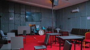 A room with a red carpet, multiple chairs, tables, a drum set, and a television screen on the wall. - Quinta Nova Hotel (Kigali)