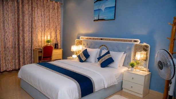 A neatly made bed with a headboard, bedside lamps, a nightstand, a fan, and slippers on the floor. - Quinta Nova Hotel (Kigali)