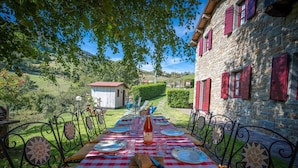 Un área de comedor al aire libre con mantel a cuadros, sillas de metal y un edificio de piedra con contraventanas rojas al fondo.
